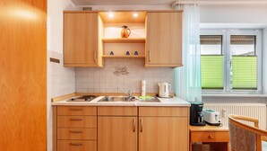 A compact kitchen with wooden cabinets, a sink, a countertop with a kettle, and a window with blinds. - Room 'Doppelzimmer 3 - Behindertengerecht' with Shared Terrace and Wi-Fi (Bad Bellingen)
