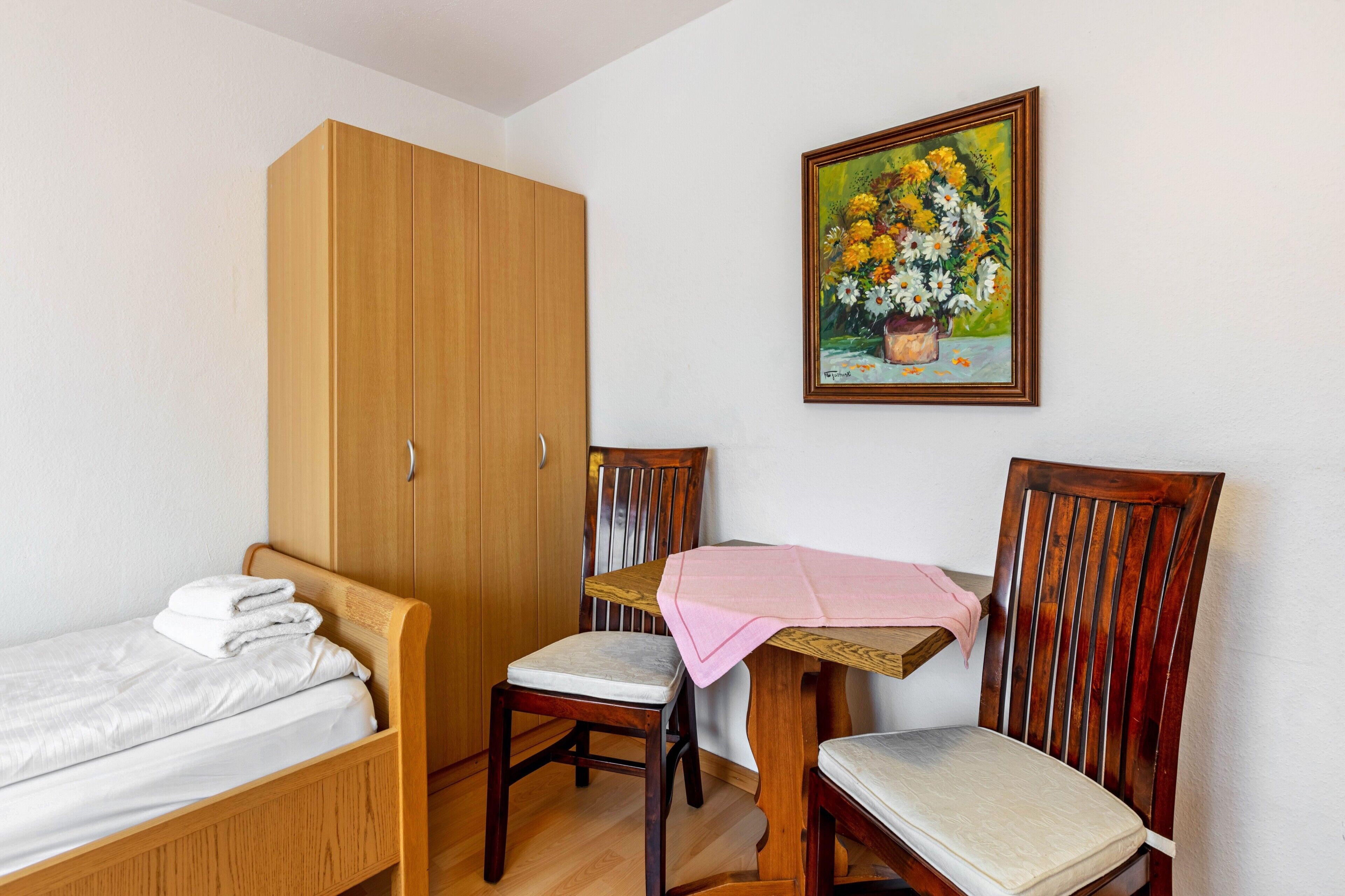 A room with a bed, a wooden wardrobe, a wooden chair, and a table with a pink cloth.