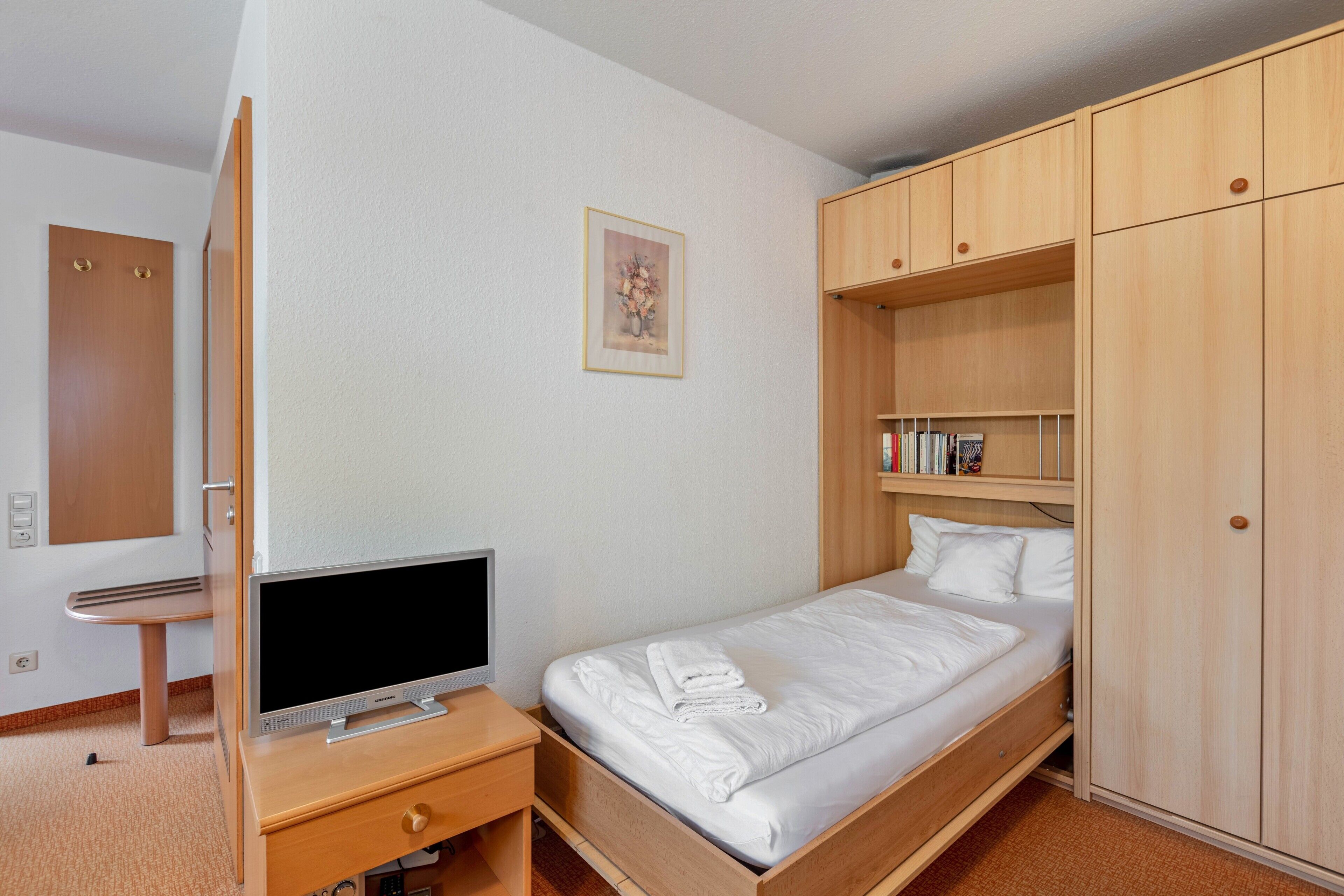 A bedroom with a bed, a TV on a wooden stand, a wardrobe, and a small desk with a chair.