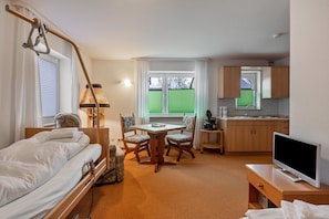 Una habitación de hotel con cama, una mesa con sillas, televisor, kitchenette y ventana con persianas.