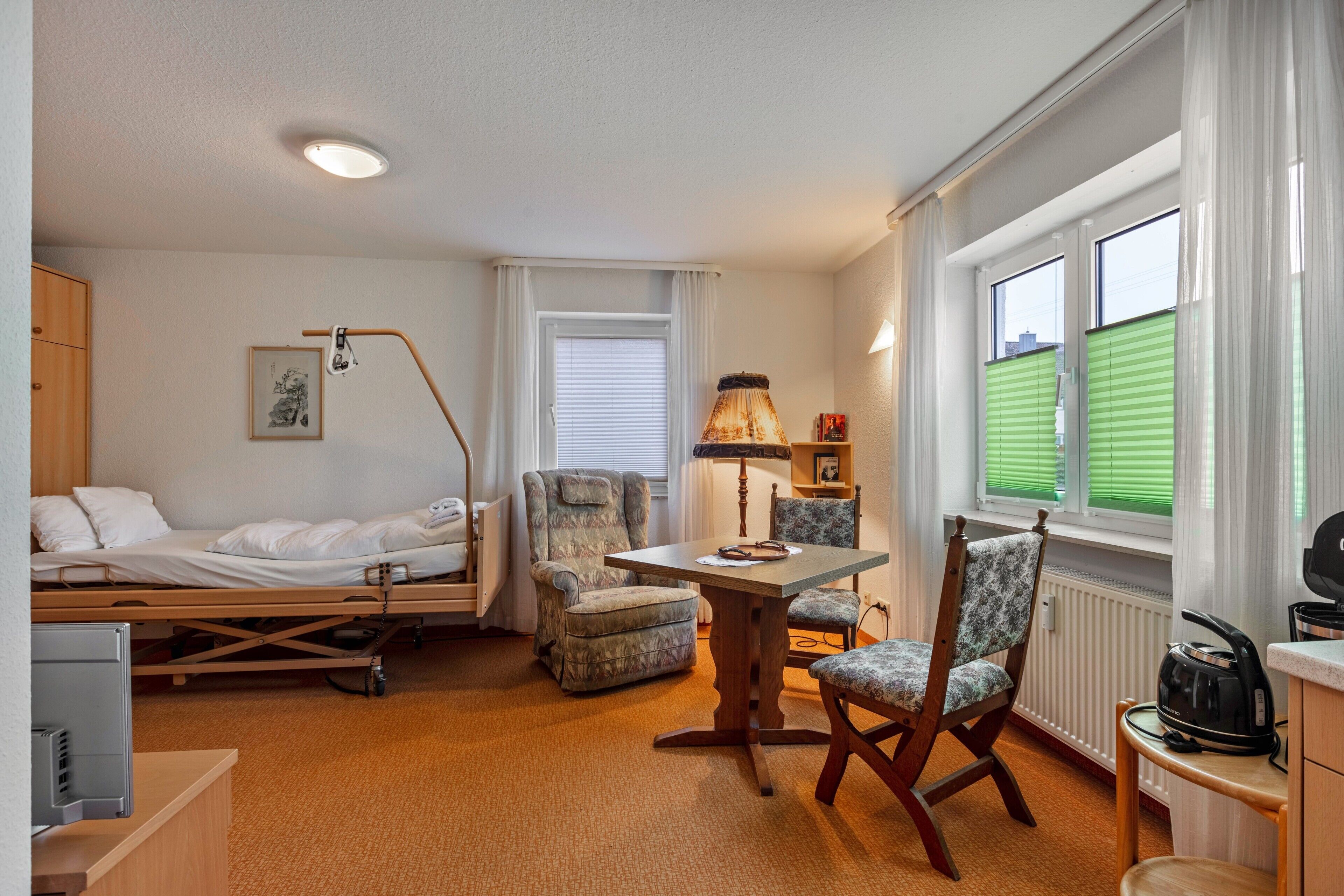 Une chambre d’hôtel comprenant un lit, un bureau, deux chaises, une lampe, un radiateur et une fenêtre avec des stores.