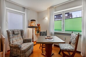 Una habitación con una mesa de comedor, dos sillones estampados, una lámpara de pie y vista a un campo verde a través de una ventana.