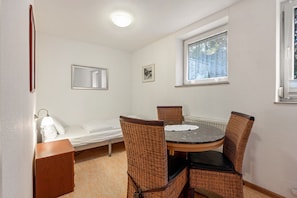Una habitación pequeña y ordenada con una cama individual, una mesa redonda de comedor con sillas, un ventanal con vistas y un espejo fijado a la pared.