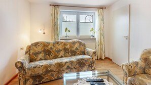Una sala de estar con un sofá estampado, una mesa de centro de vidrio, una ventana con cortinas y una puerta.