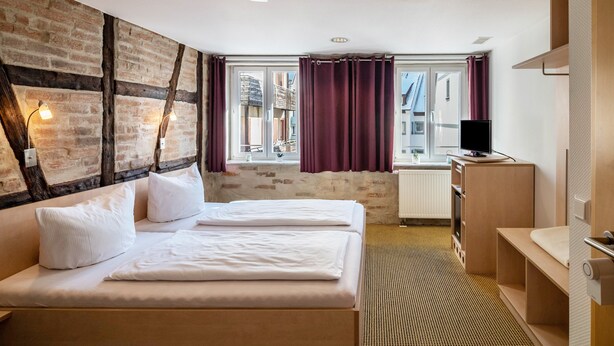 Een hotelkamer met een bakstenen muur, twee bedden, een bureau, een televisie en een raam met gordijnen.