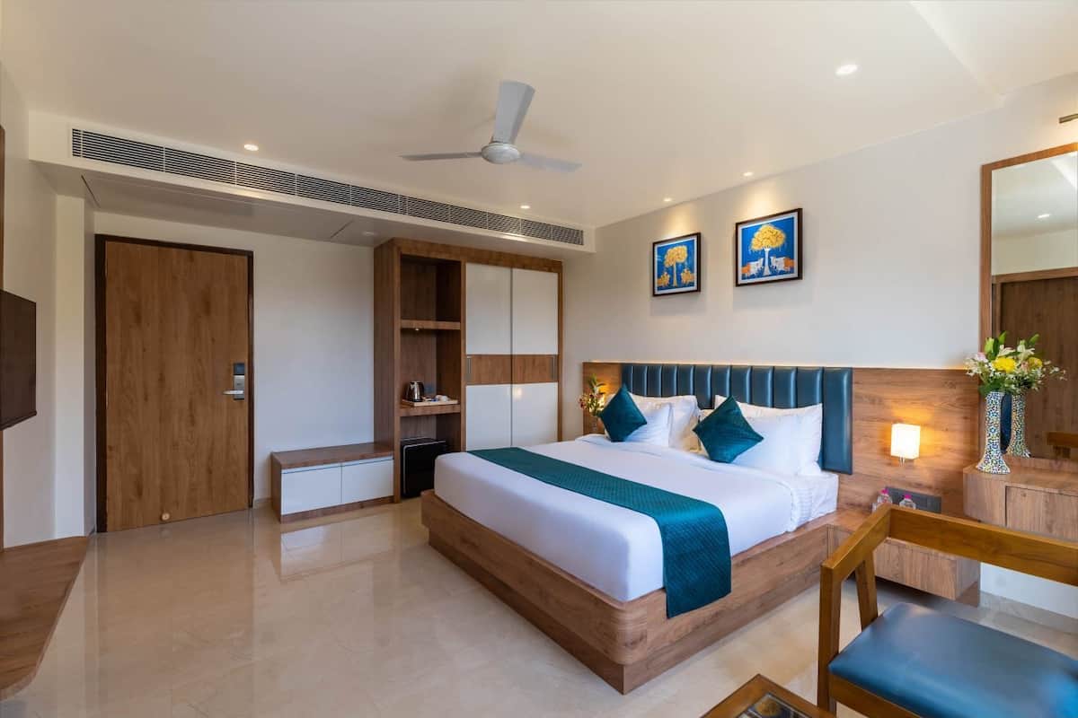 Une chambre d’hôtel moderne avec un grand lit, une tête de lit en bois, un ventilateur de plafond, une table de chevet, un miroir et des œuvres d’art accrochées au mur.