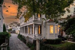 Ett vitt hus i kolonialstil med en veranda och ett burspråk, omgivet av grönska och en gång belagd med kullersten.