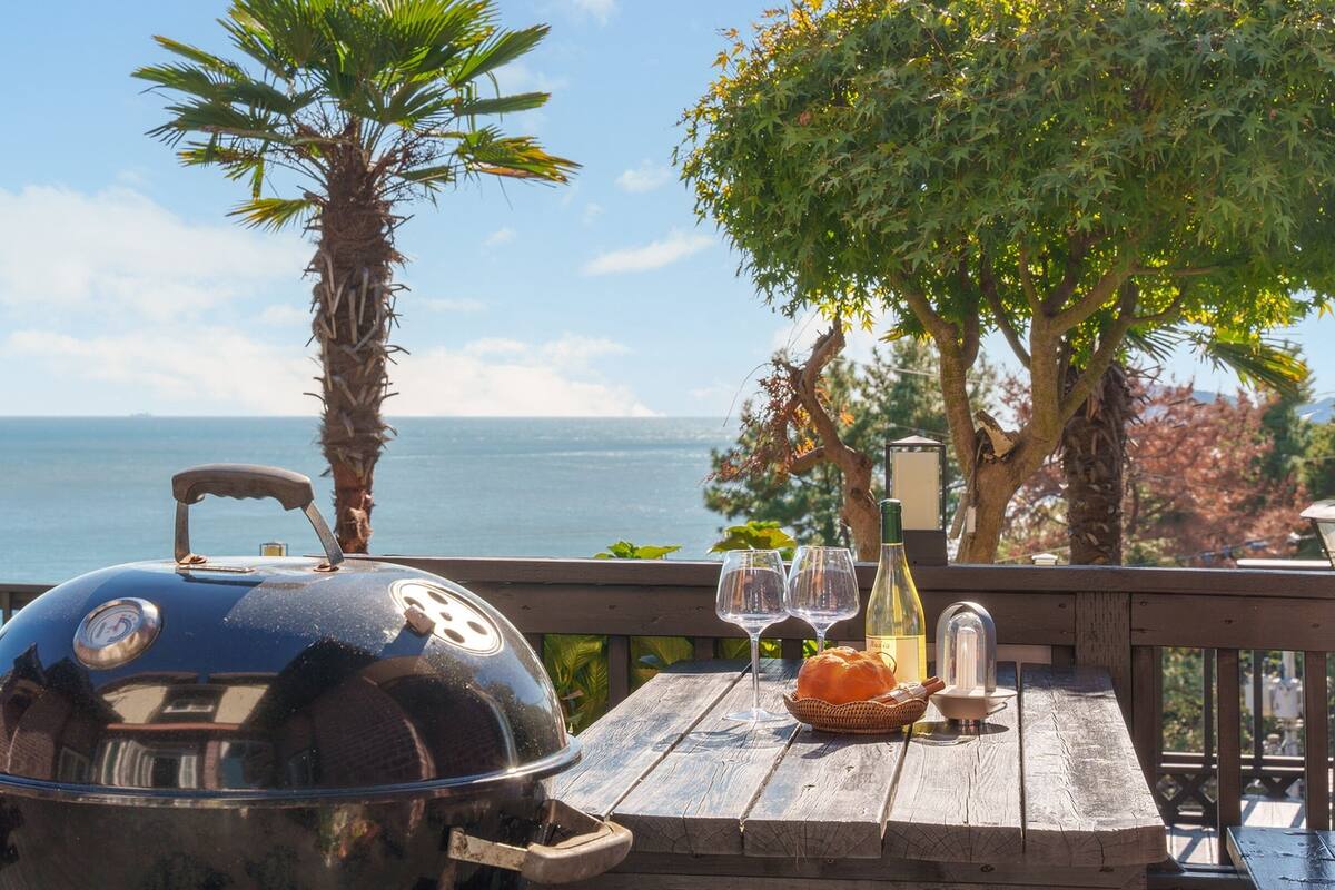 A barbecue grill on a wooden table with a view of the sea and trees.