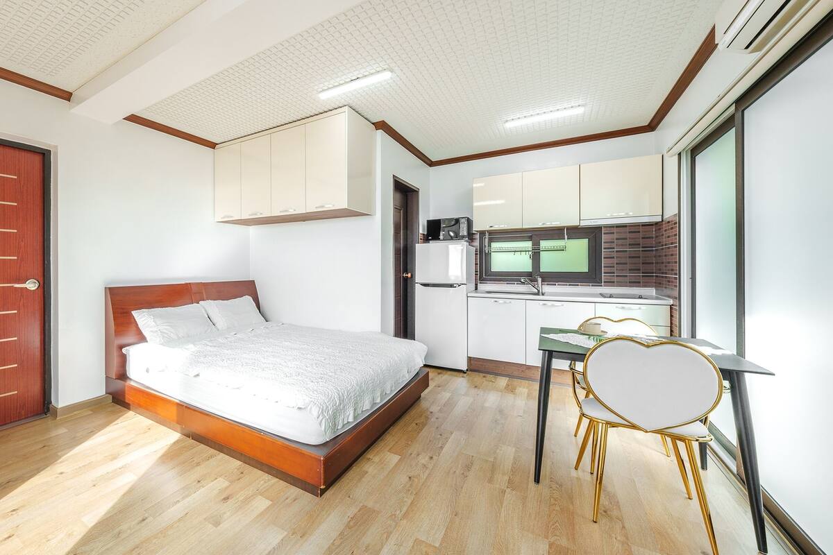 A bedroom with a wooden bed, a desk with a chair, and a kitchen area with a refrigerator.