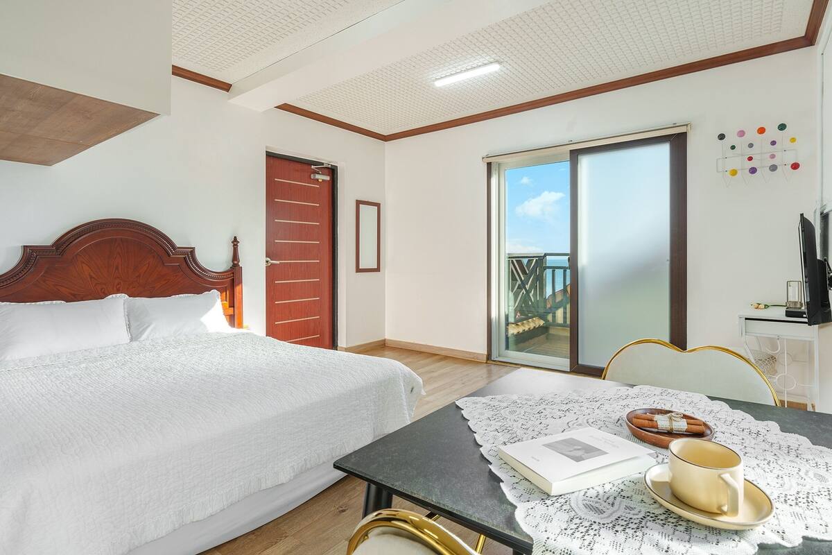 A hotel room with a bed, a table with a book and a cup, a TV, and a balcony with a view.
