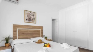 Een netjes ingerichte hotelkamer met een bed met houten hoofdeinde, witte lakens, een fruitschaal, een hoed en handdoeken op het bed.