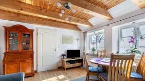 Une pièce avec des poutres en bois, un téléviseur, une table à manger avec des chaises, un buffet et une fenêtre avec une plante.