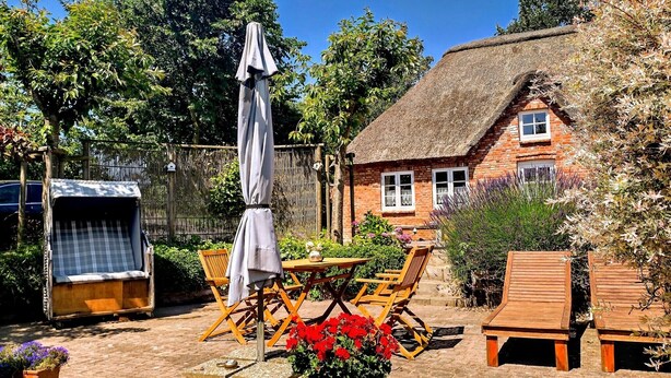 Une maison en briques avec un toit de chaume, une terrasse avec des meubles en bois et des plantes en pot.