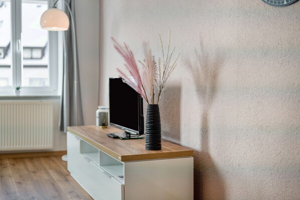 Une chambre moderne avec un bureau en bois, un vase noir contenant des plantes séchées et un téléviseur posé sur une commode blanche.