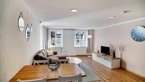 Ein modernes Wohnzimmer mit Esstisch, weißen Stühlen, einer Couch und einem Fernseher auf einem weißen Schrank, sowie einer großen Wanduhr.
