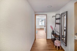 A hallway with a wooden floor, a pink stool, a mirror with a decorative frame, and a radiator in the background.