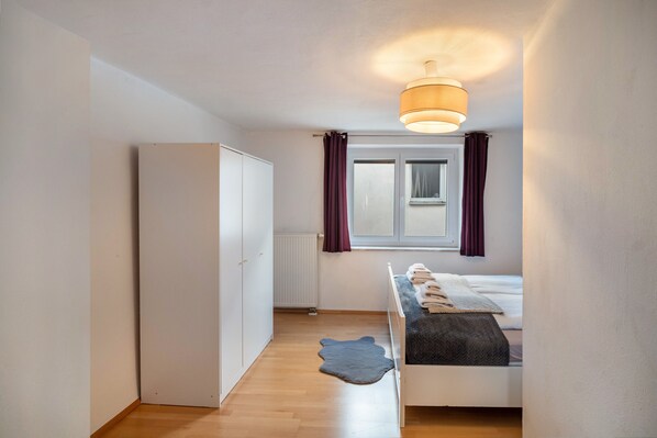 A bedroom with a bed, a wardrobe, a radiator, a window with curtains, and a ceiling light.