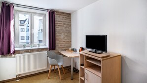 A room with a window, a radiator, a desk with a chair, a television, and a brick wall. - Single Room 52 with Wi-Fi (Bad Waldsee)