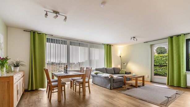 Ein Wohnzimmer mit Esstisch, Stühlen, einer Couch, einer Lampe und Blick durch ein Fenster mit Vorhängen.
