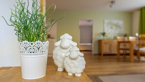 Ein weißer Keramiktopf mit Grünpflanzen, eine weiße Keramik-Schafigurin und ein Holztisch in einem Raum mit grüner Wand und Holzboden.