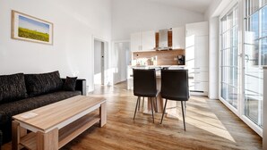 Una sala moderna con un sofá, una mesa de madera y dos sillas. Se ve una cocina al fondo con armarios blancos y una ventana.