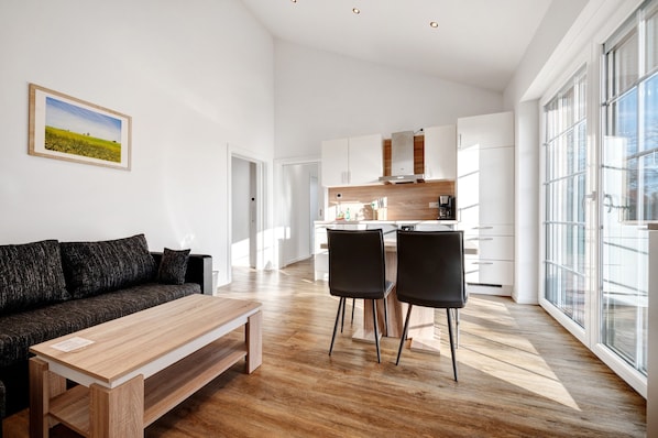 Una sala moderna con un sofá, una mesa de madera y dos sillas. Se ve una cocina al fondo con armarios blancos y una ventana.