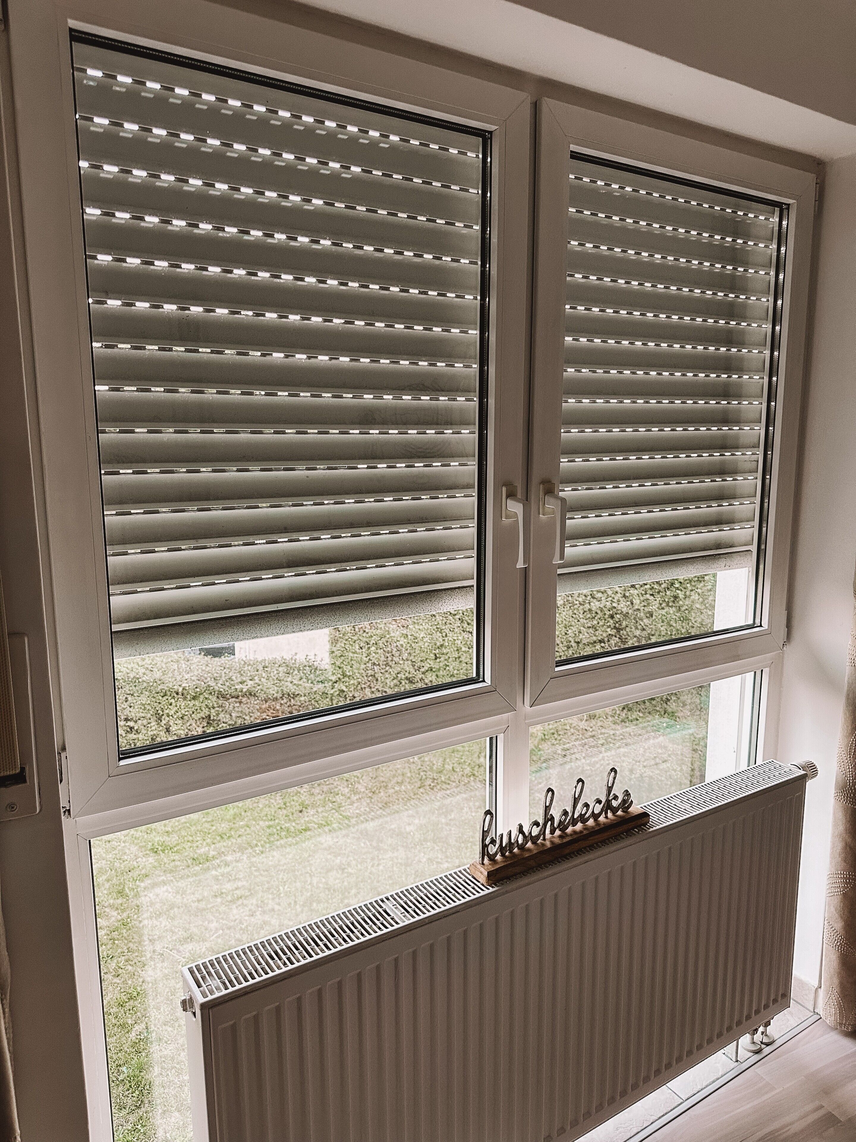 A room with two windows featuring horizontal blinds, a radiator below, and a small wooden sign with text.