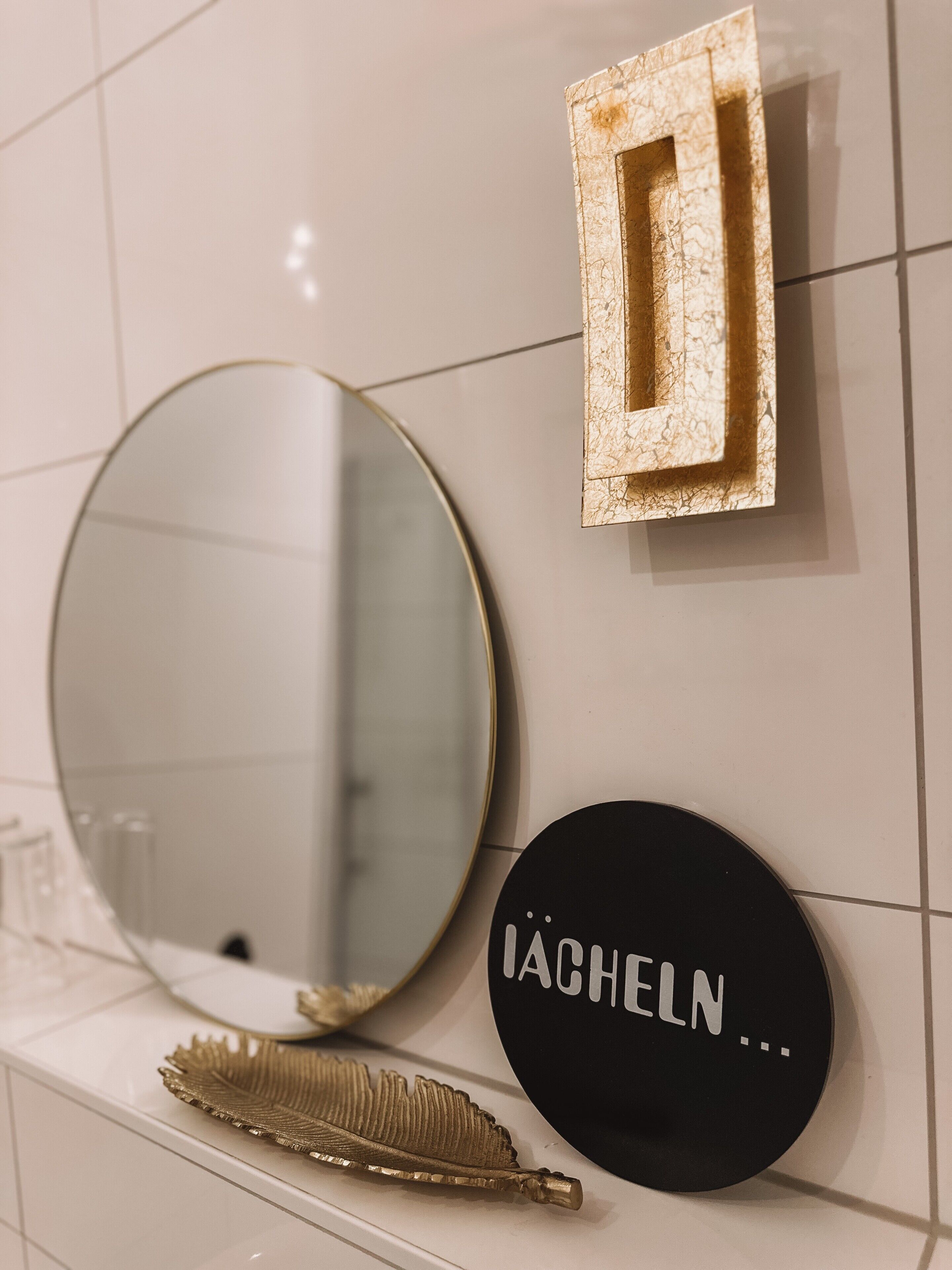 Ein Badezimmer mit einem runden Spiegel, einem rechteckigen Spiegel mit goldener Rahmen und einem schwarzen, runden Schild mit Text.