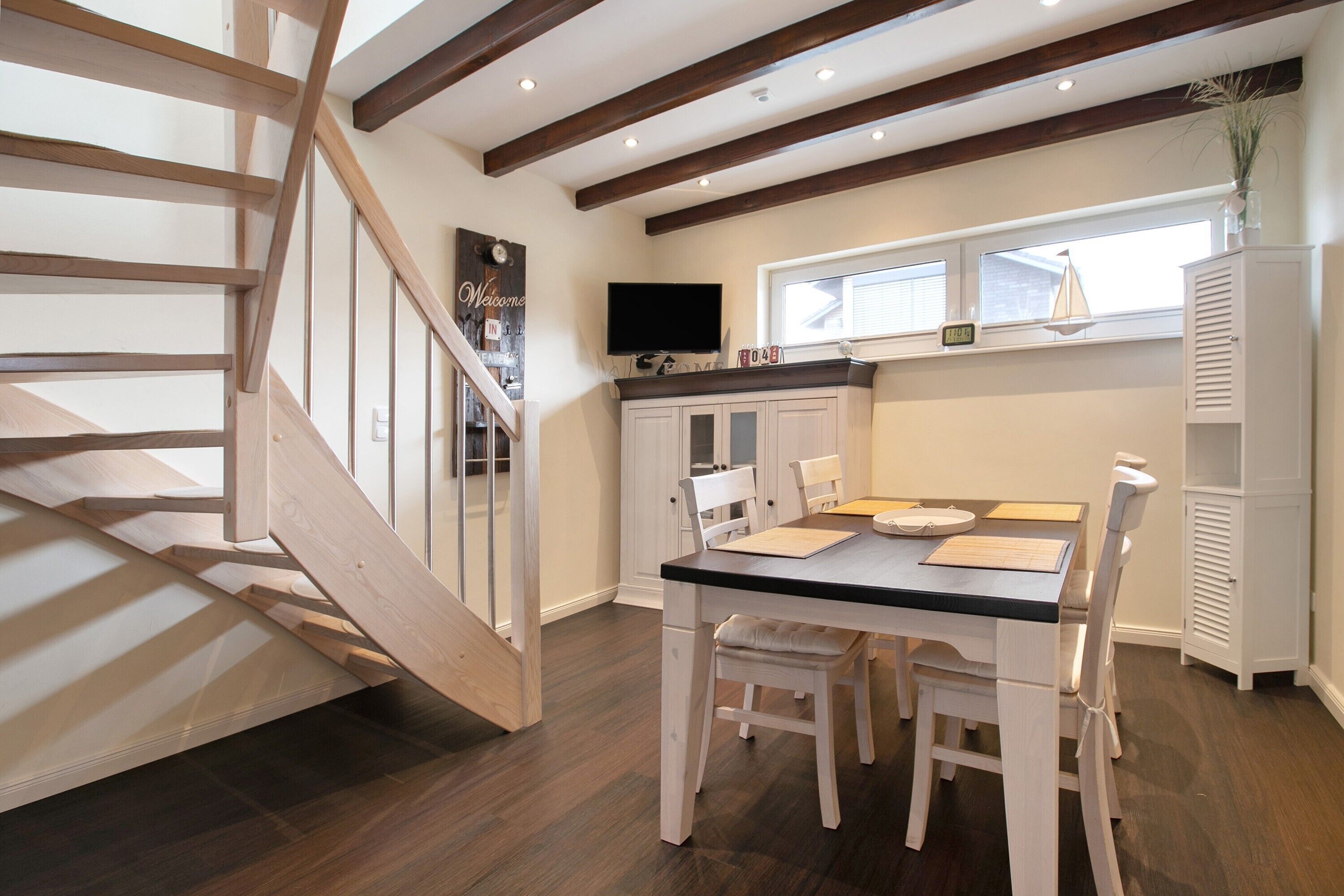 A dining area with a table and chairs, a staircase, a TV mounted on the wall, and a window with blinds.