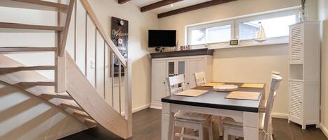 A dining area with a table and chairs, a staircase, a TV mounted on the wall, and a window with blinds.