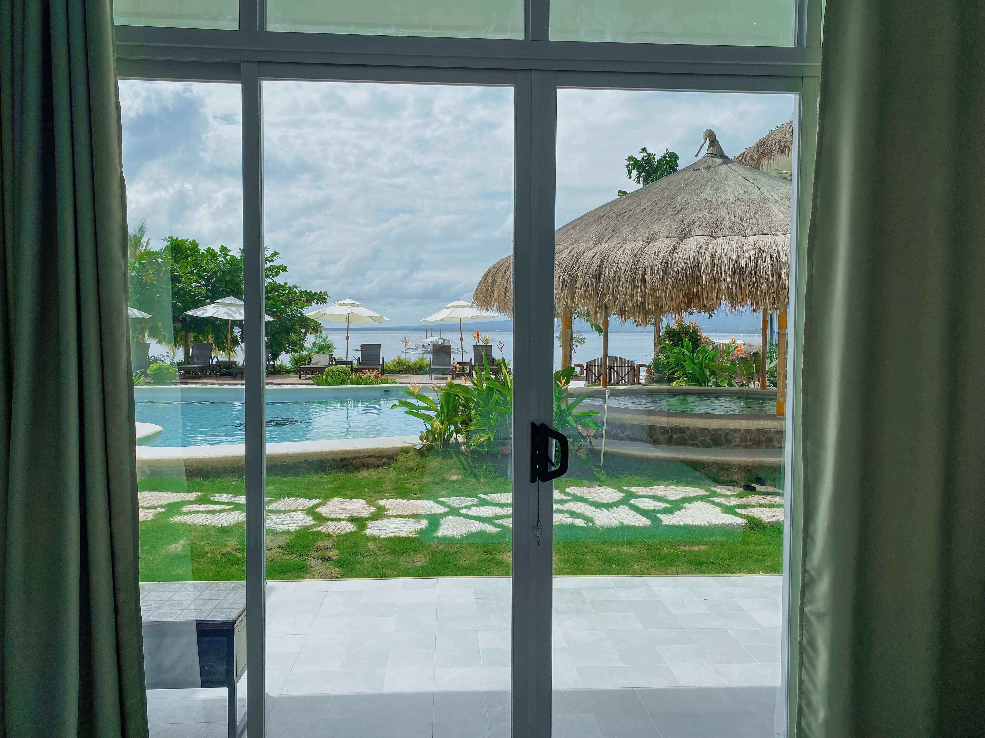 A view of a swimming pool with lounge chairs and umbrellas, a thatched-roof structure, and a grassy area with a patterned lawn.