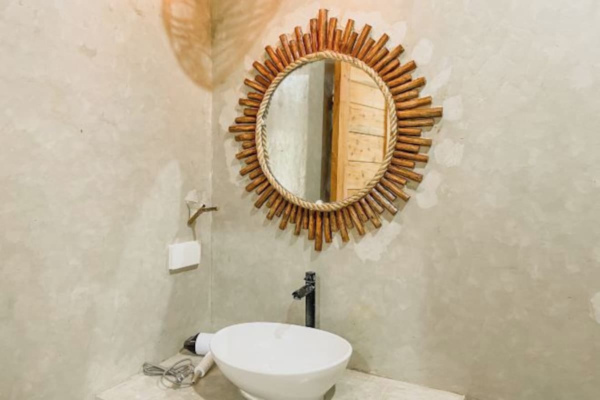 Une salle de bain avec un miroir rond encadré de bois, un lavabo blanc et un distributeur de serviettes.