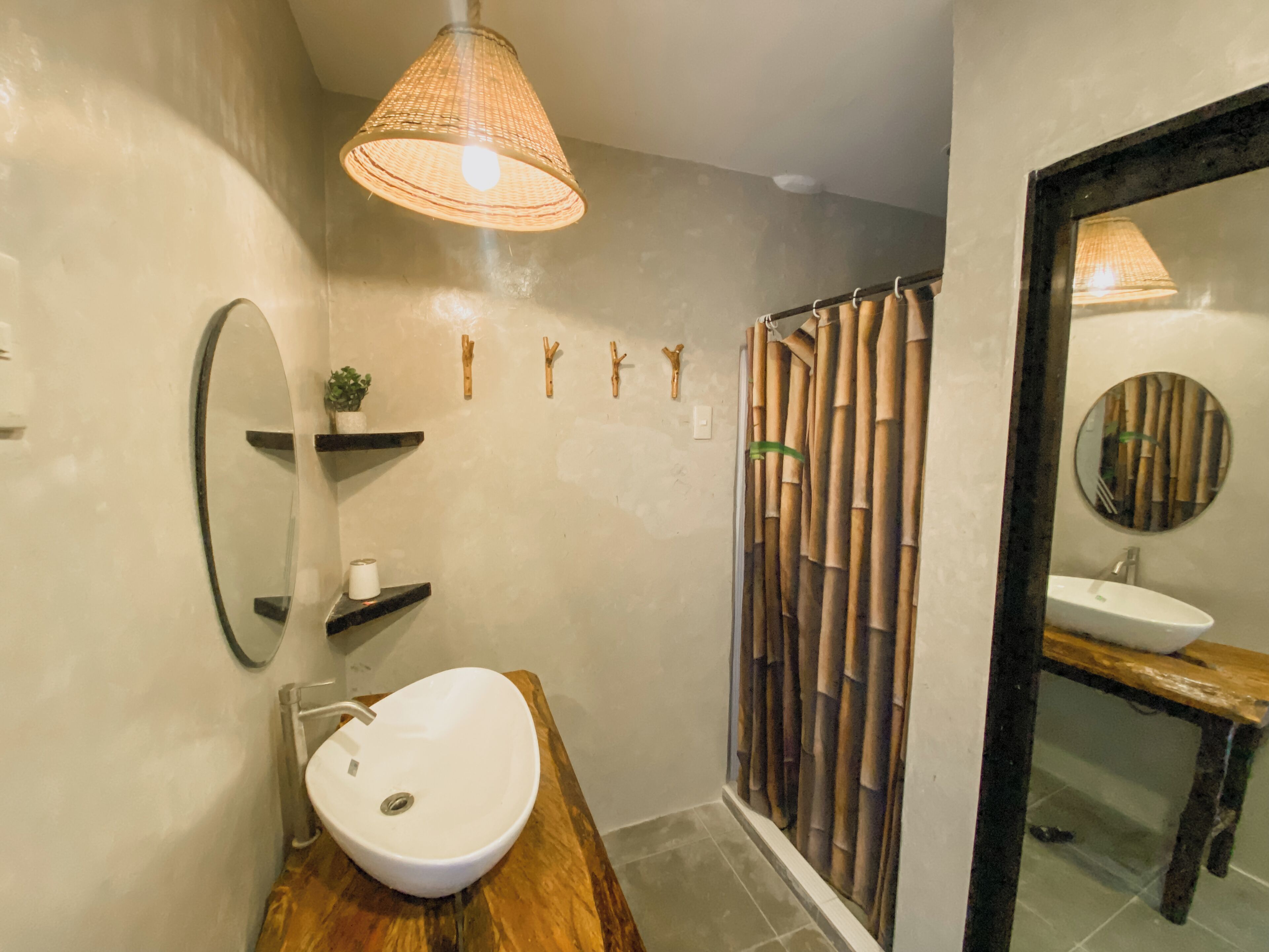 Un baño con tocador de madera, un lavamanos blanco, un espejo, una lámpara colgante, cortinas de bambú y una planta pequeña en un estante.