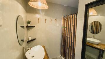 Un baño con tocador de madera, un lavamanos blanco, un espejo, una lámpara colgante, cortinas de bambú y una planta pequeña en un estante.