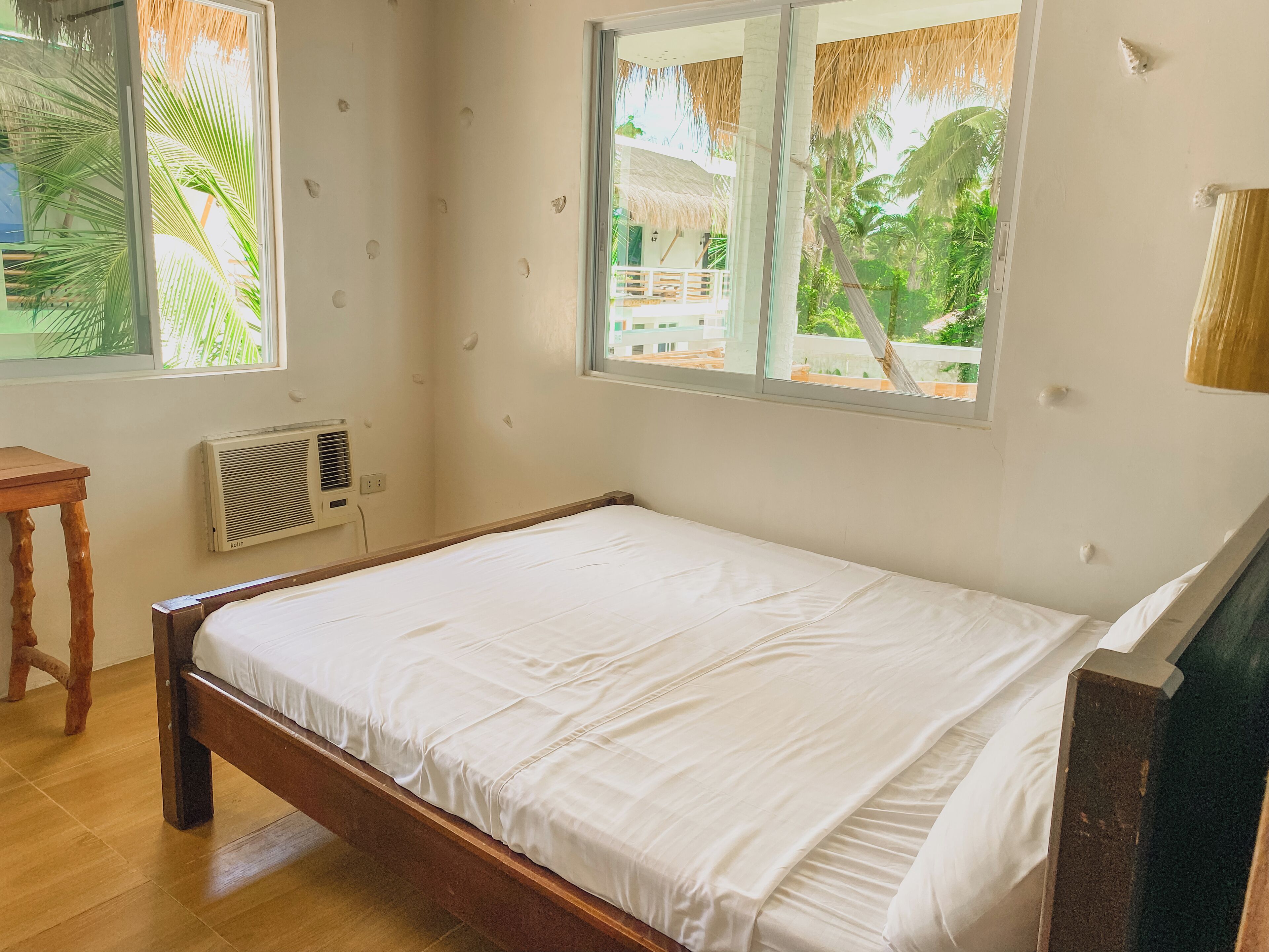 Un dormitorio con cama de madera, una ventana con vista a palmeras y una mesita.