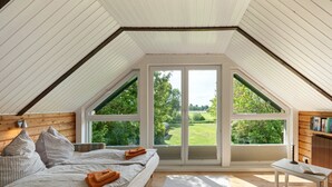 A bedroom with a sloped ceiling, large windows, a bed with pillows, a bedside table, and a bookshelf.
