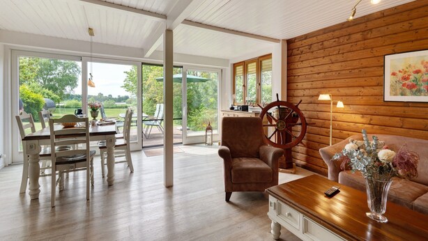 A spacious room with a wooden ceiling, a dining area with a table and chairs, a leather armchair, a sofa, and a vase with flowers.