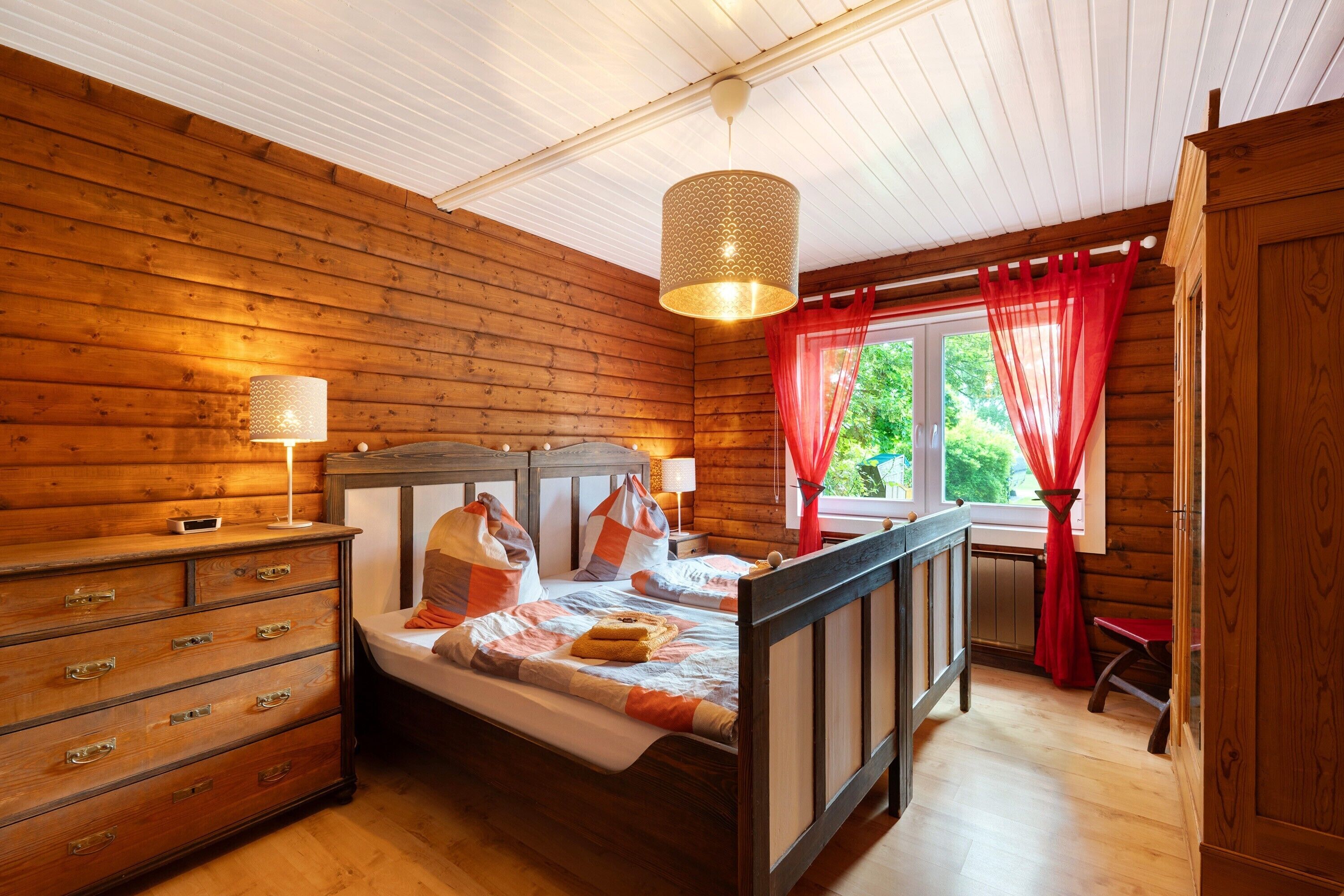 Ein Schlafzimmer einer Holzhütte mit Bett, Schminktisch und einem Fenster mit roten Vorhängen.
