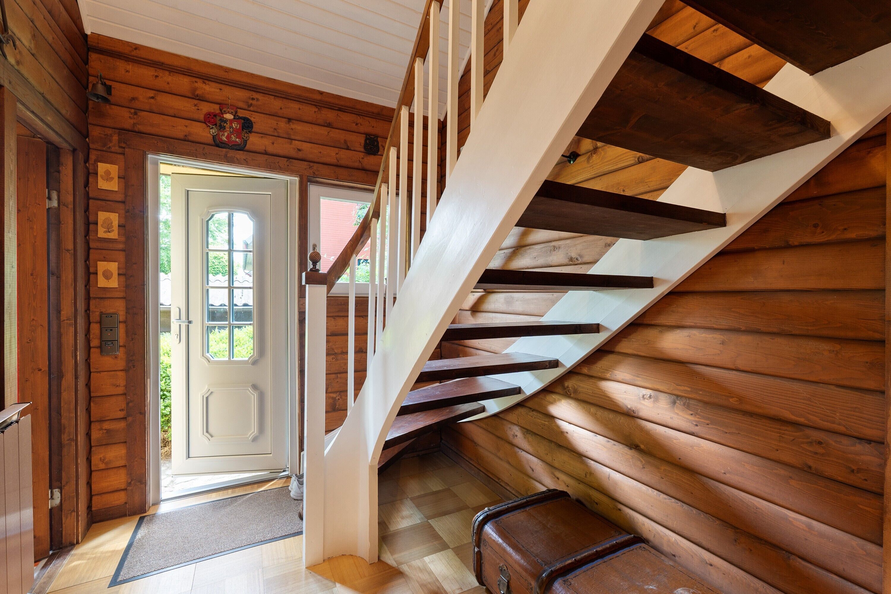 Interior em madeira com escada, porta para o exterior e mala no chão.