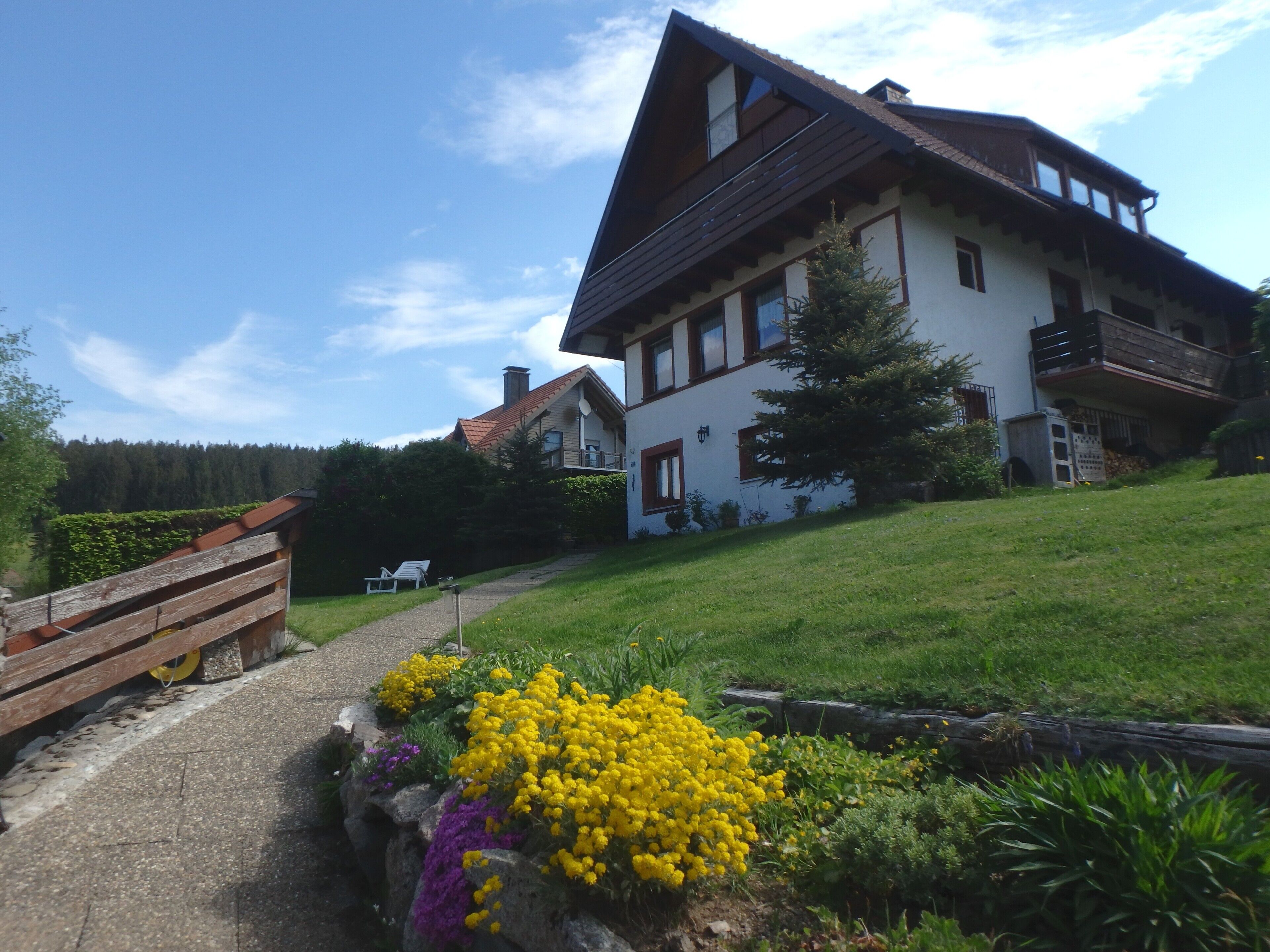 A two-story house with a sloped roof, a balcony, and a garden with yellow flowers.