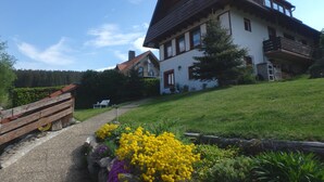 Une maison à deux étages, dotée d’un toit en pente, d’un balcon et d’un jardin où poussent des fleurs jaunes.