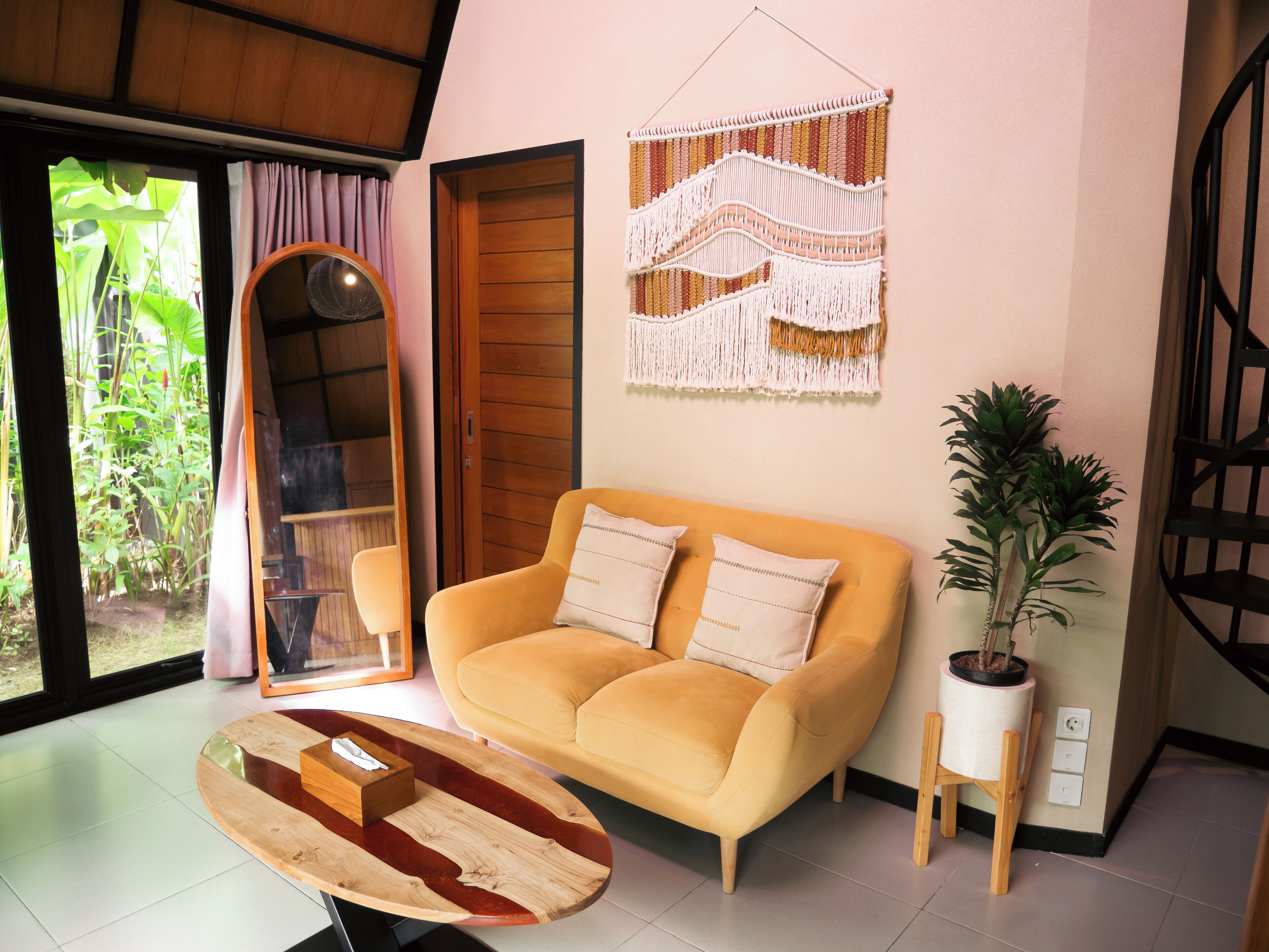 A cozy living room with a yellow sofa, a wooden coffee table, a potted plant, and a macrame wall hanging.