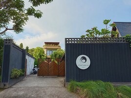 Una entrada porticada a una villa con un logotipo circular, un edificio de techo plano y vegetación circundante.