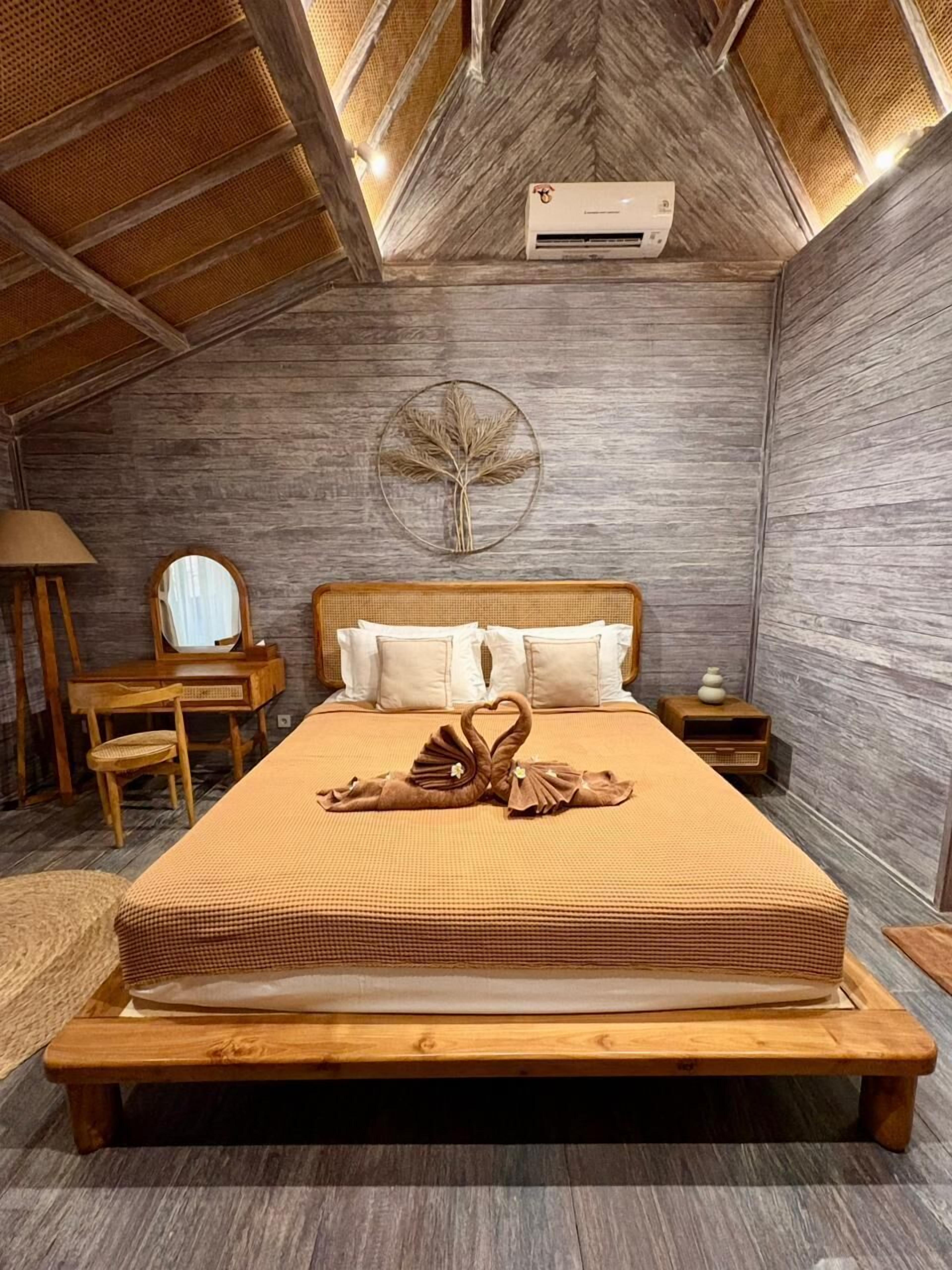 A bedroom with a wooden bed, a round mirror, a bedside table, a wall-mounted fan, and a ceiling fan.