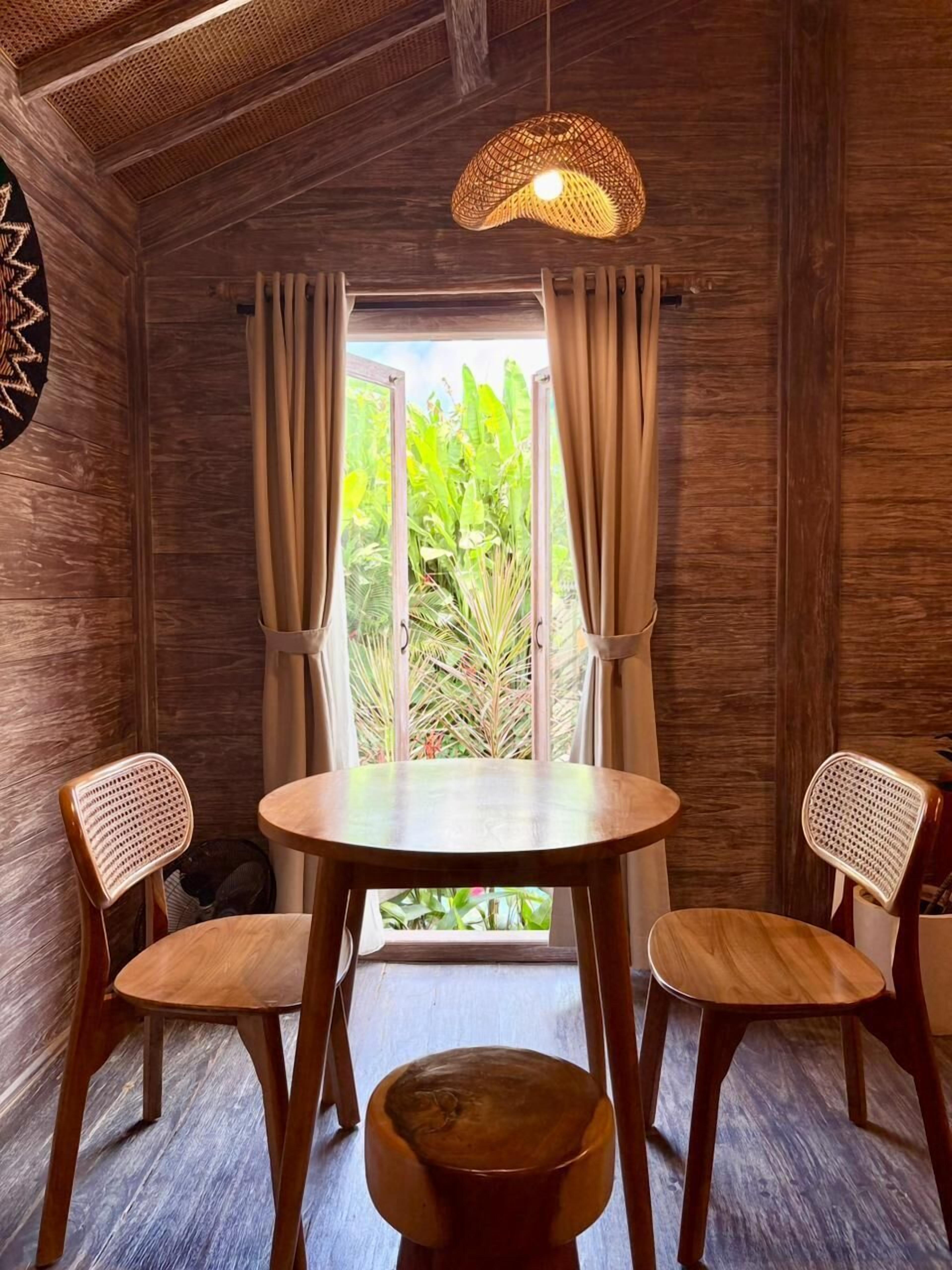 Une pièce avec une table ronde en bois, deux chaises en osier, un luminaire suspendu en fibres tressées et une vue sur la verdure par une fenêtre ornée de rideaux.