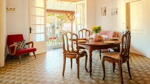 A dining area with a wooden table, chairs, and a chandelier. There is a red chair and a white chair with a red cushion. A vase with flowers is on the table.