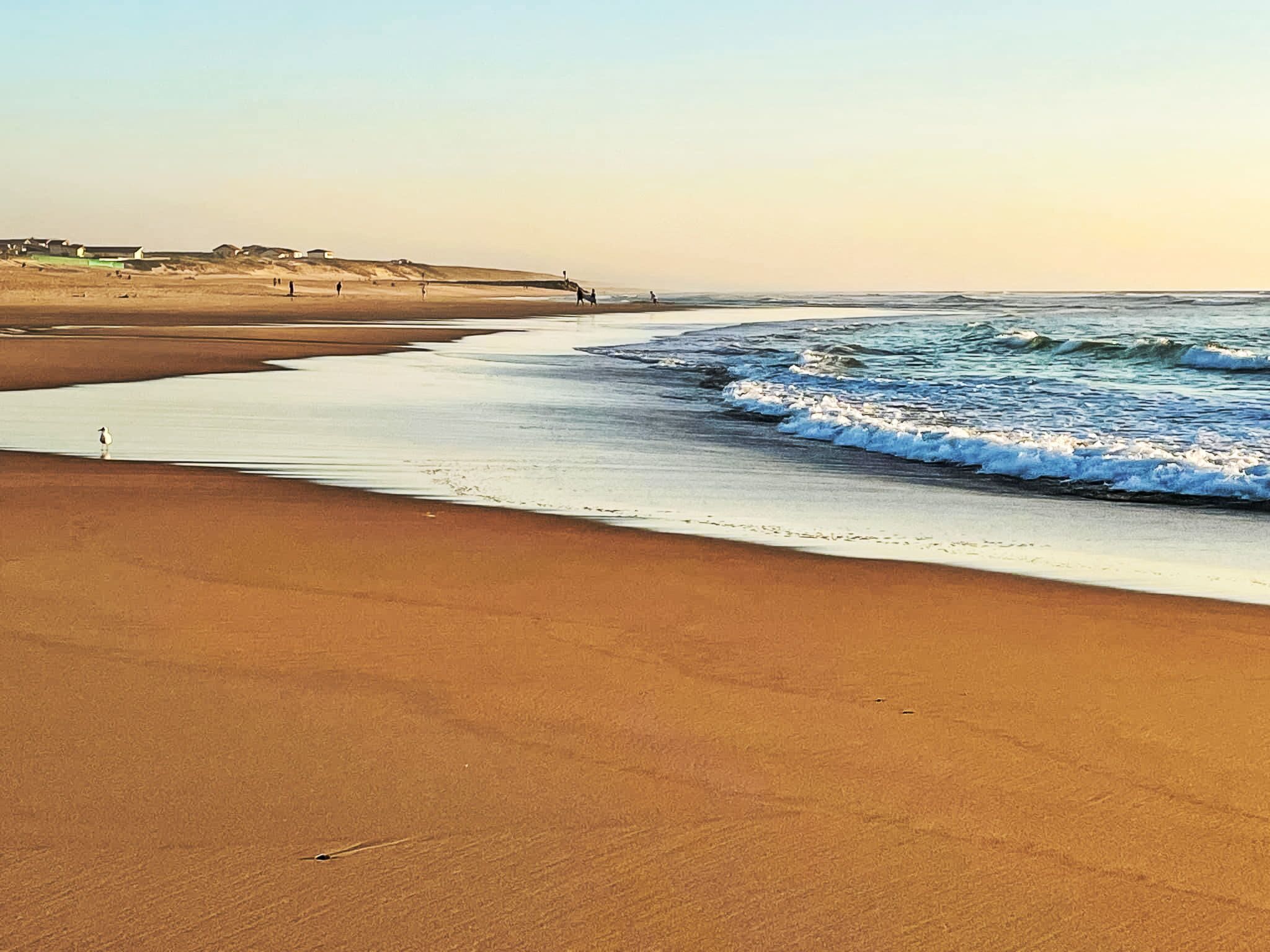 A beach with a wide sandy shore, gentle waves, and a few people walking along the water's edge.