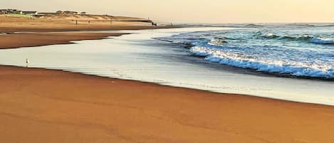 Een strand met een breed zandstrand, rustige golven en enkele mensen die langs de waterlijn lopen.
