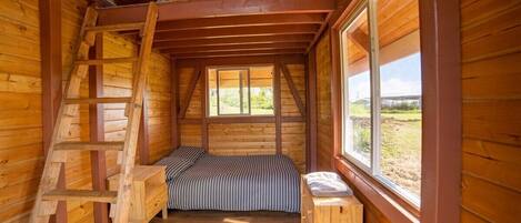 A cozy attic bedroom with a loft bed, wooden ladder, and a window overlooking a grassy field.