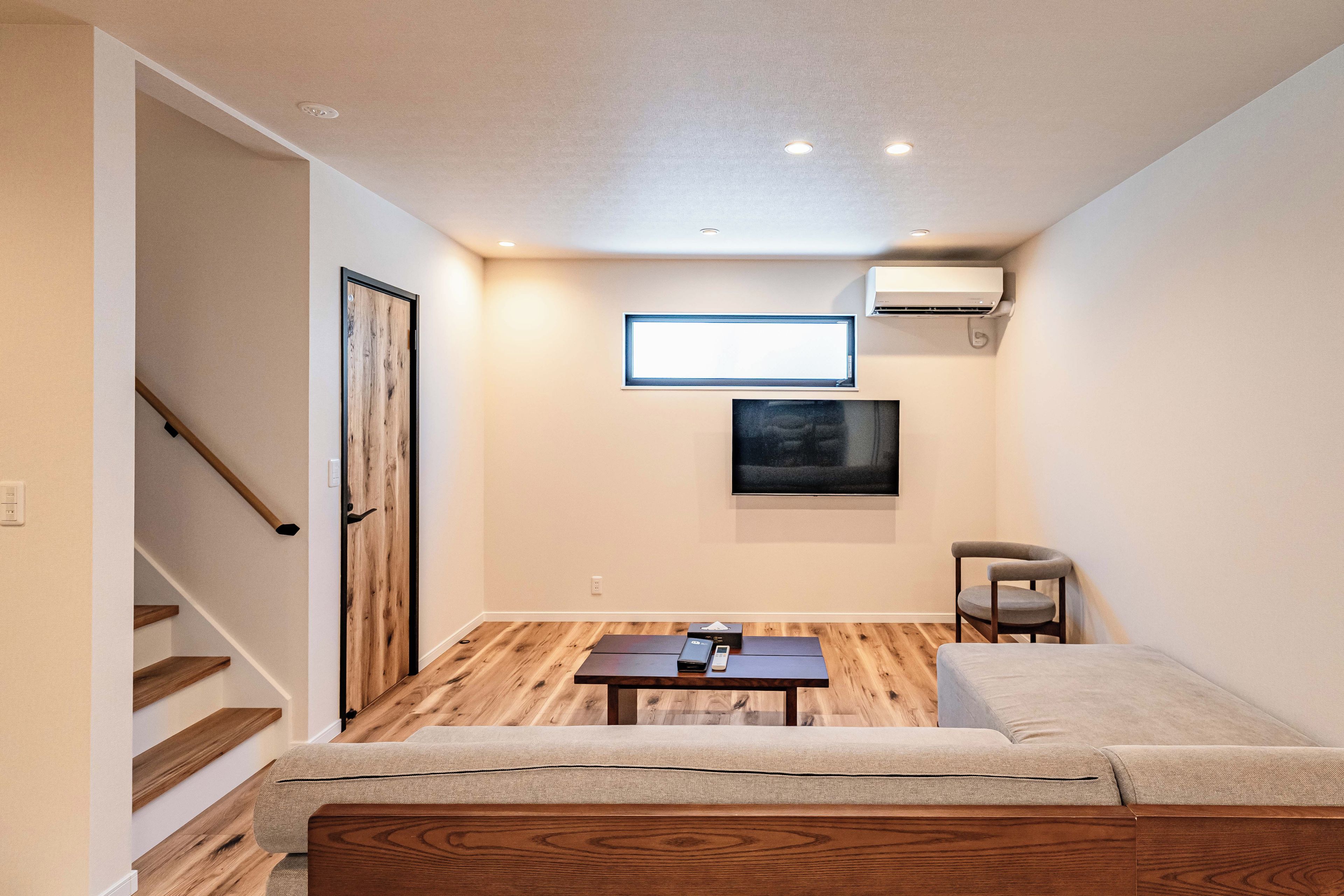 A compact living space with a sofa, a small table, a TV mounted on the wall, and a staircase leading to another area.